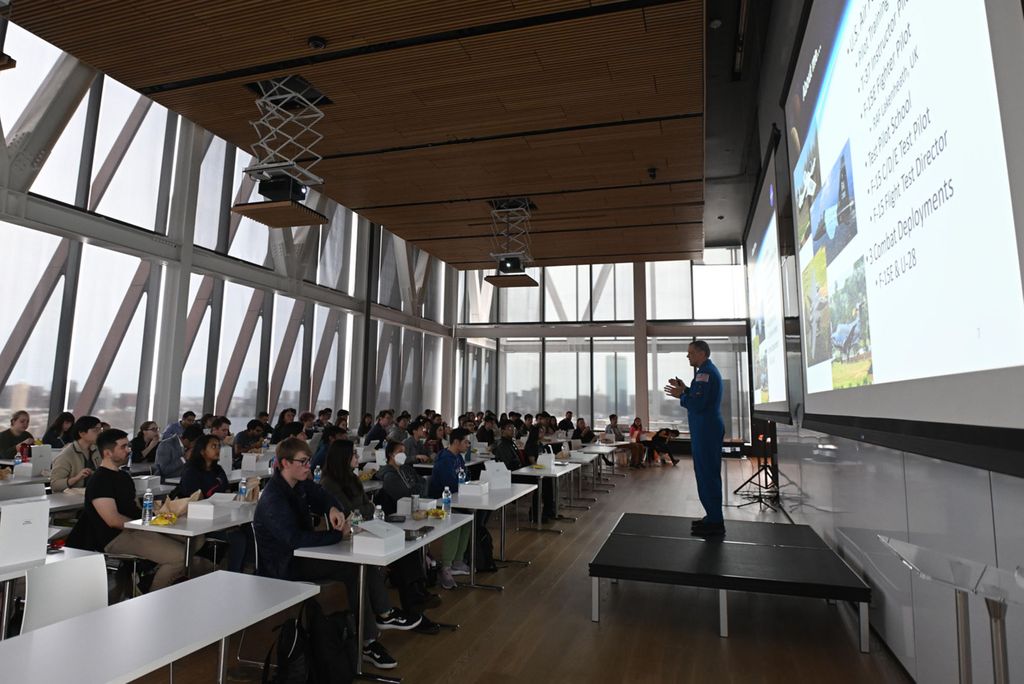 Photo: Astronaut Bob Hines (ENG’97) speaks with ENG students April 7 on the 17th floor of CCDS. A man in a blue NASA astronaut jumpsuit stands on a makeshift stage at the front of a large room as he speaks to a large audience of students sitting and watching.