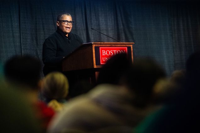 Photo: A black woman wearing glasses stands at a podium with a red placard reading "Boston University" in front of a crowd of people