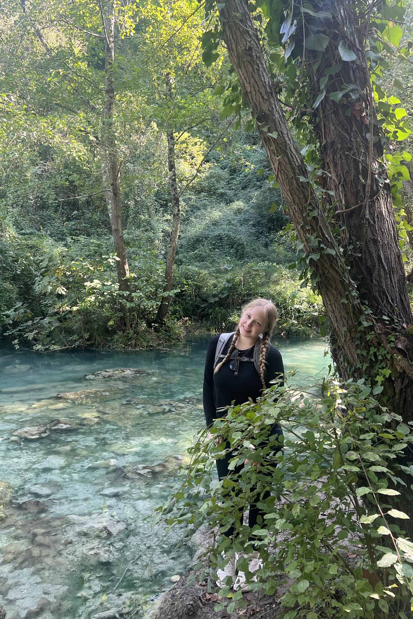 Photo: Katherine, a young white woman, stands in a forest space next to a churring body of water. A bush is in front of her.