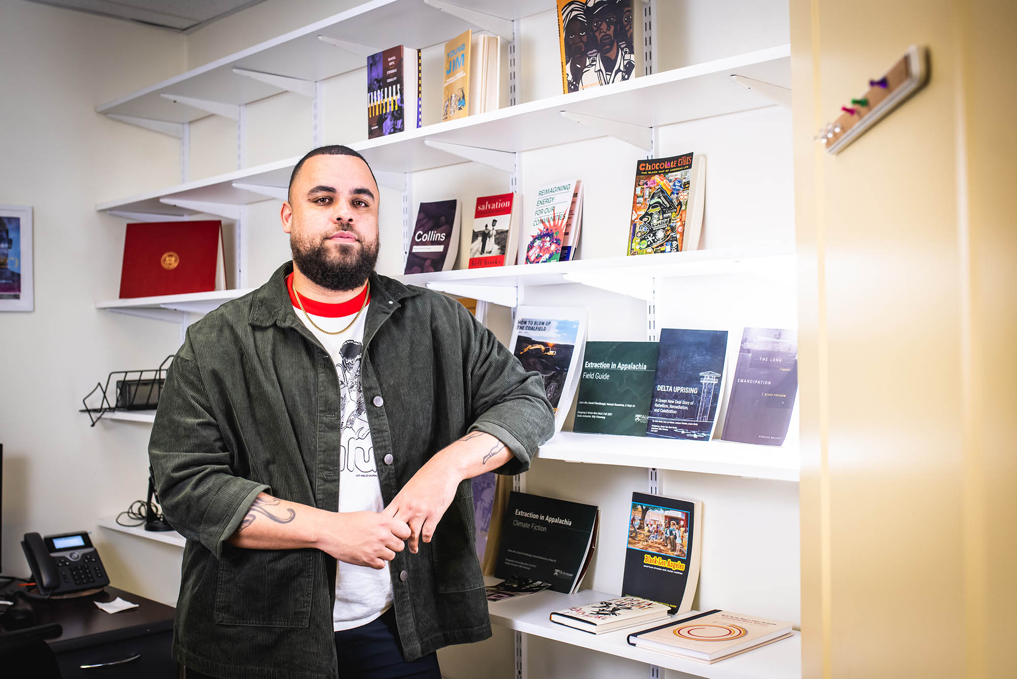 Photo: Darien Alexander Williams, a man with short, dark hair and a beard poses with his hands clasped, arm resting on a wall shelf. He wears a green button up and a white shirt with a red collar.