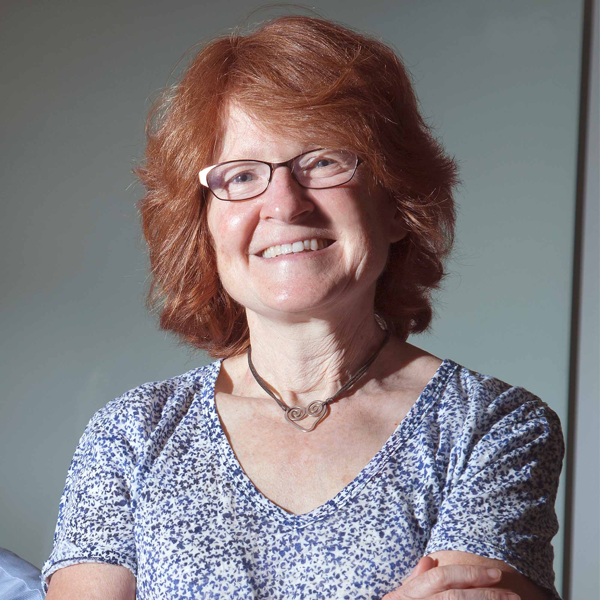 Photo: A woman with red hair and glasses smiles for a studio portrait on a grey background.