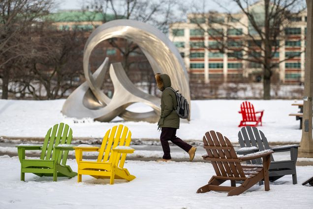 Photo: A student walks across a snow covered campus in January. A silver statue stands in the background and in the foreground, colorful chairs. The student is obscured by their winter coat.