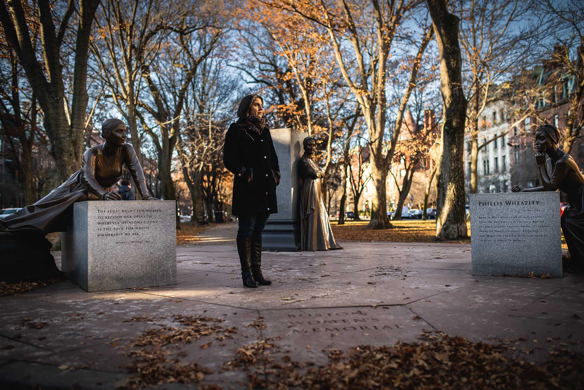 Photo: A women stands outside in the cold with three statues. There are dead trees behind her, indicating it is winter.