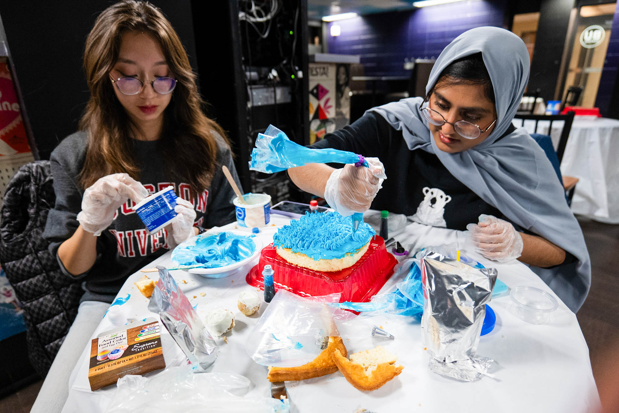 Photo: Two woman diligently decorate cakes for a BU event. On the left, a woman with dark, long hair. On the right, a woman with a blue-gray hijab.