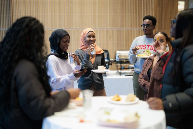 Photo: A group of college students gather around a table with appetizers and drinks. One of the is wearing a headscarf and another is wearing glasses.