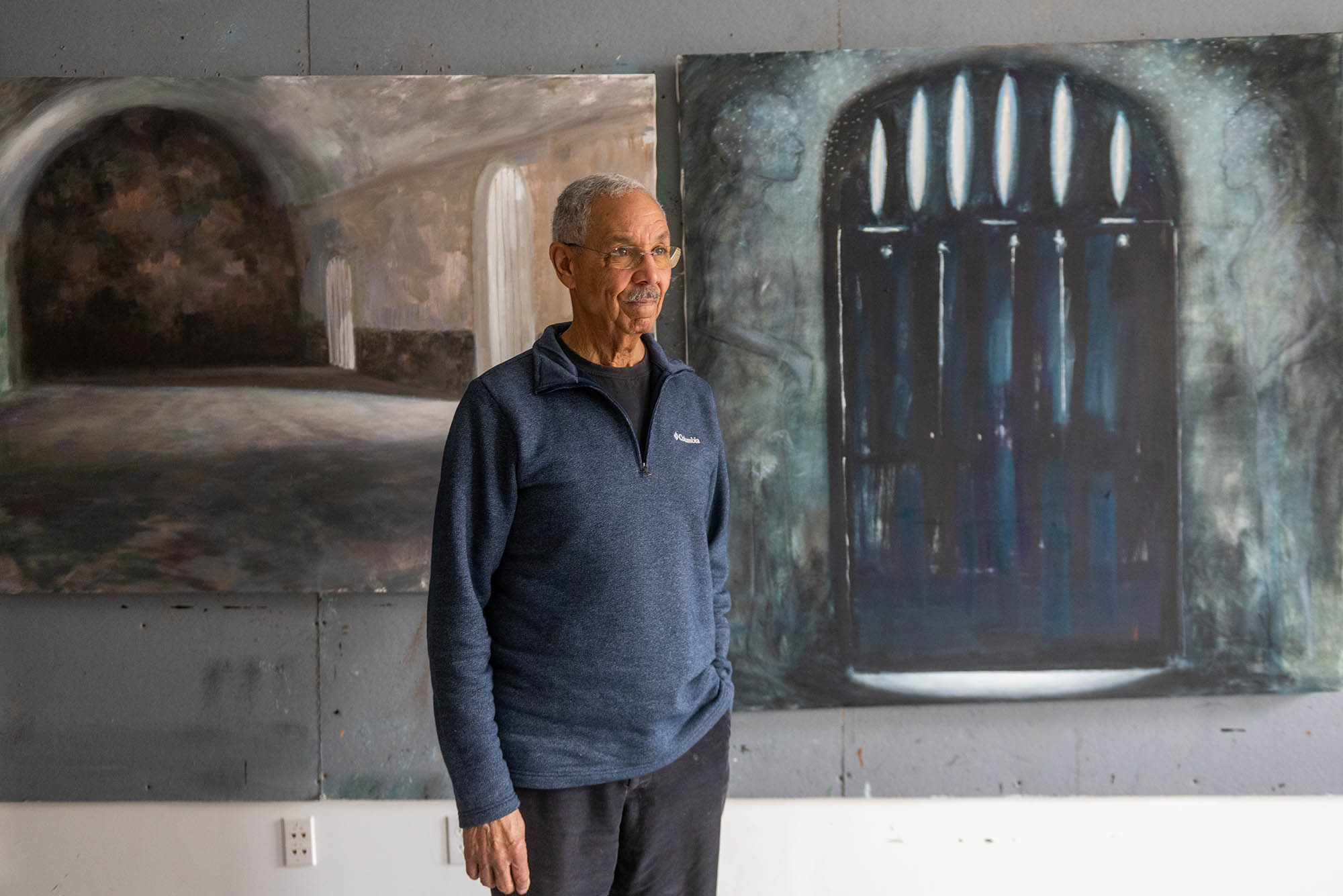 Photo: Artist Bob Freeman, a Black man with graying hair and a gray mustache, (CFA’71,’83) in his Waltham studio. The walls are used to hang his pieces, which are behind him.