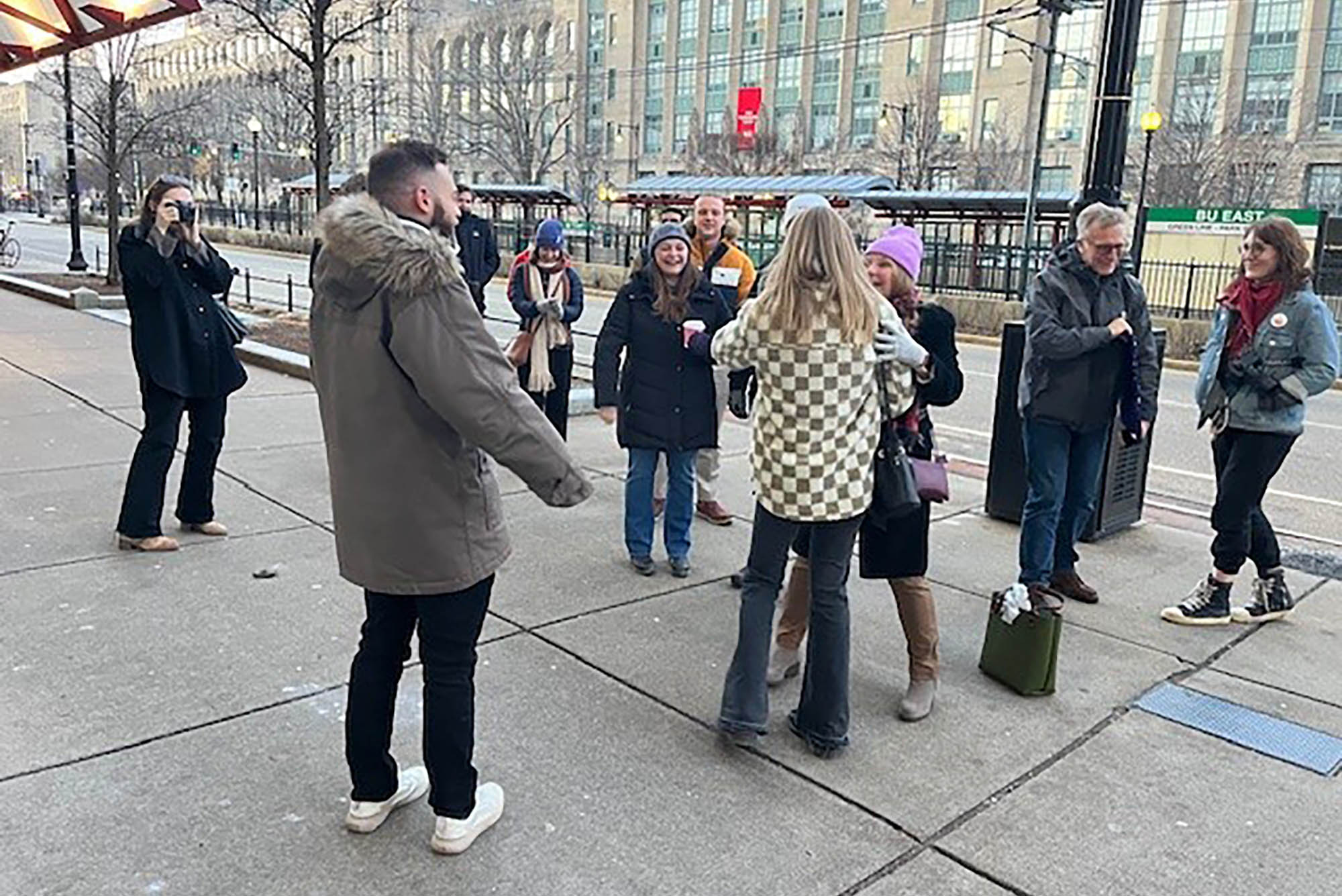 Photo: A group of people congratulate their friend on their engagement. They are wearing light coats and standing on BU's campus sidewalk.