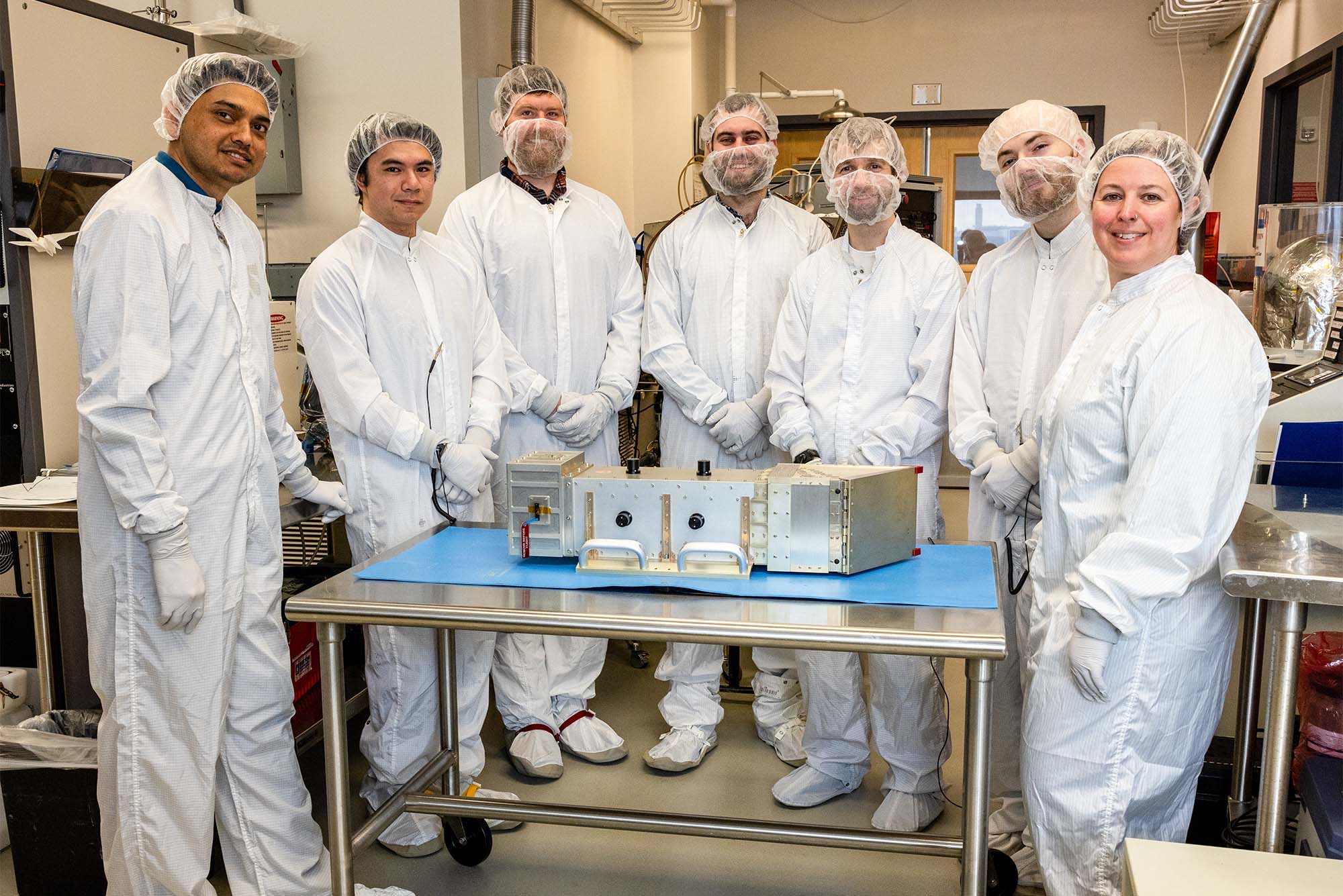 Photo: A group of scientists in white outfits and hair nets stand around a large metal machine with proud looks on their faces in a lab setting