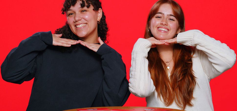 Photo: Two college students in front of a red background smile and hold their hands under their chins