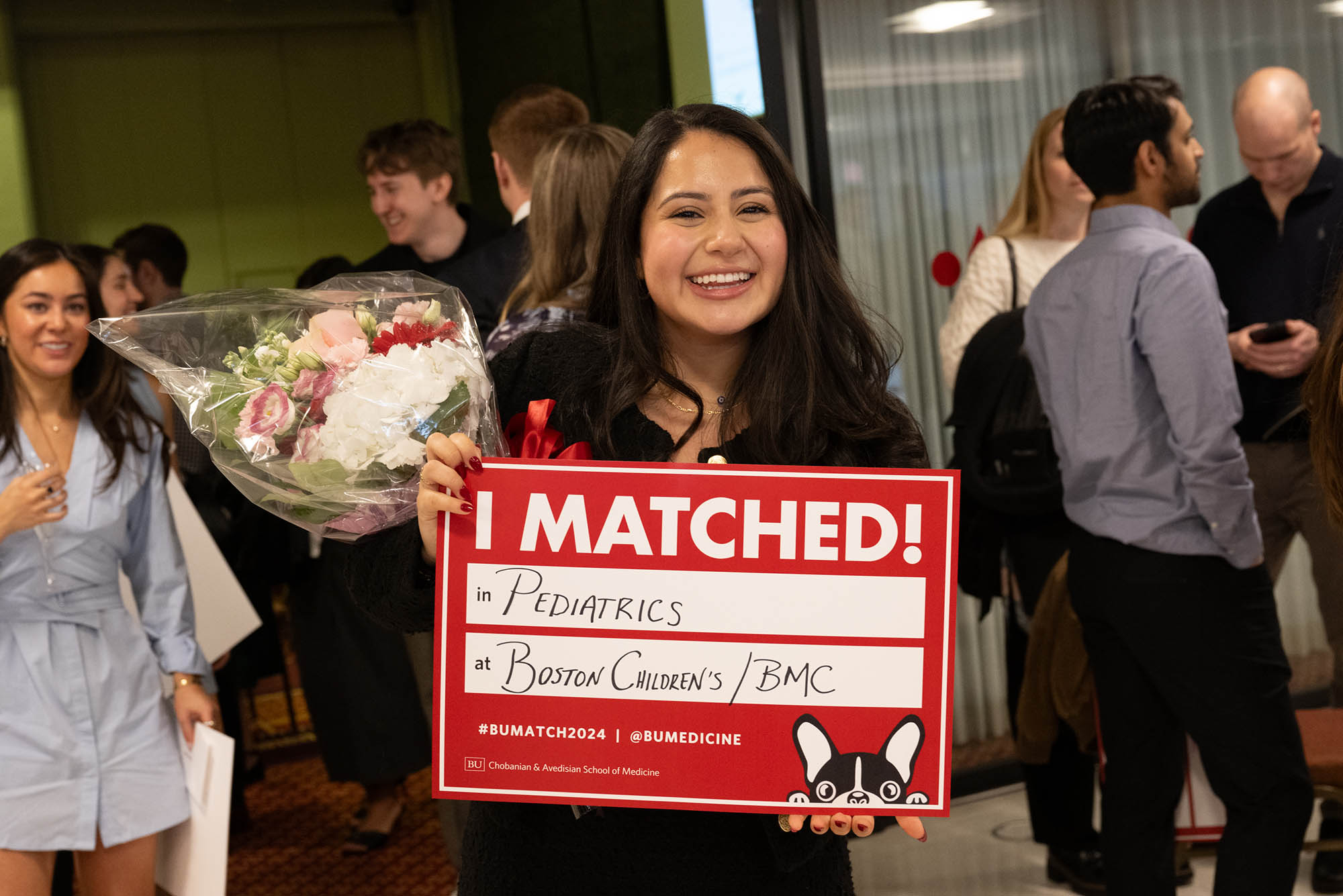 Photo: A photo of a woman smiling with a sign that says I MATCHED. She holds flowers in her hand, behind the sign.