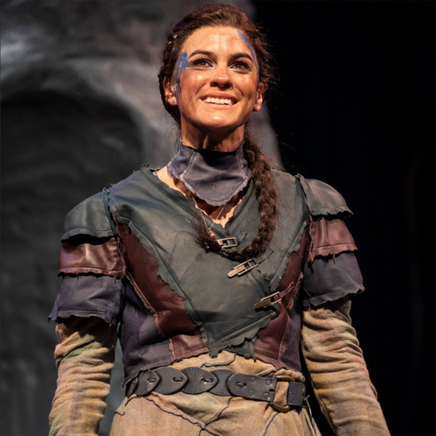 Photo: A picture of a woman on stage. She is wearing neutral, layered clothing and has a few lines of blue paint smeared on her face
