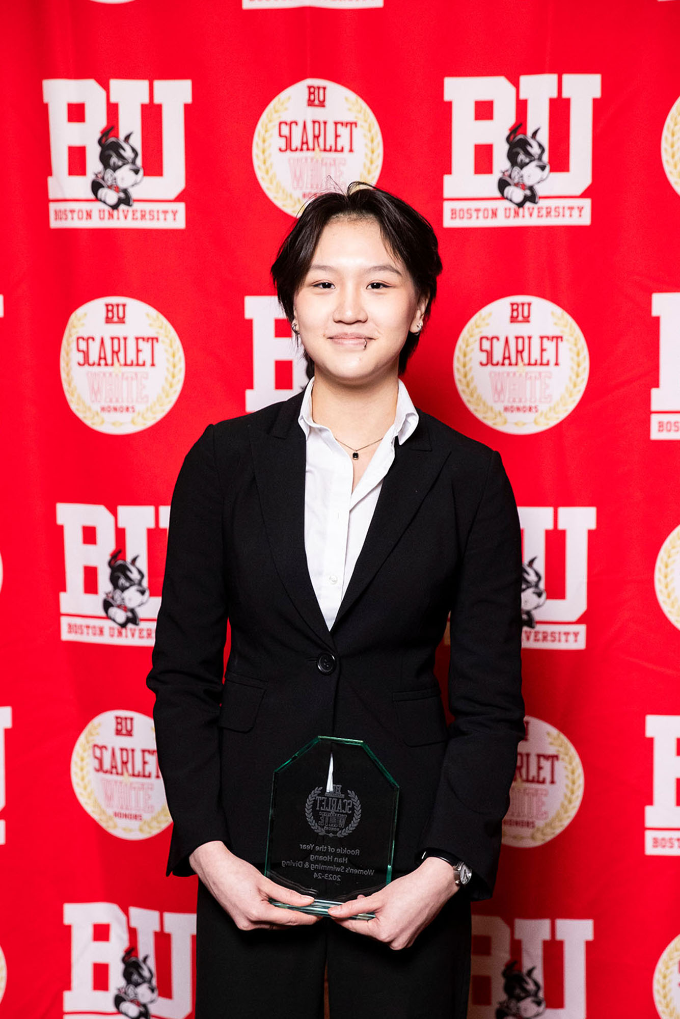 Photo: A picture of a woman in a black suit holding a clear award. There is a red background that says "BU" behind her