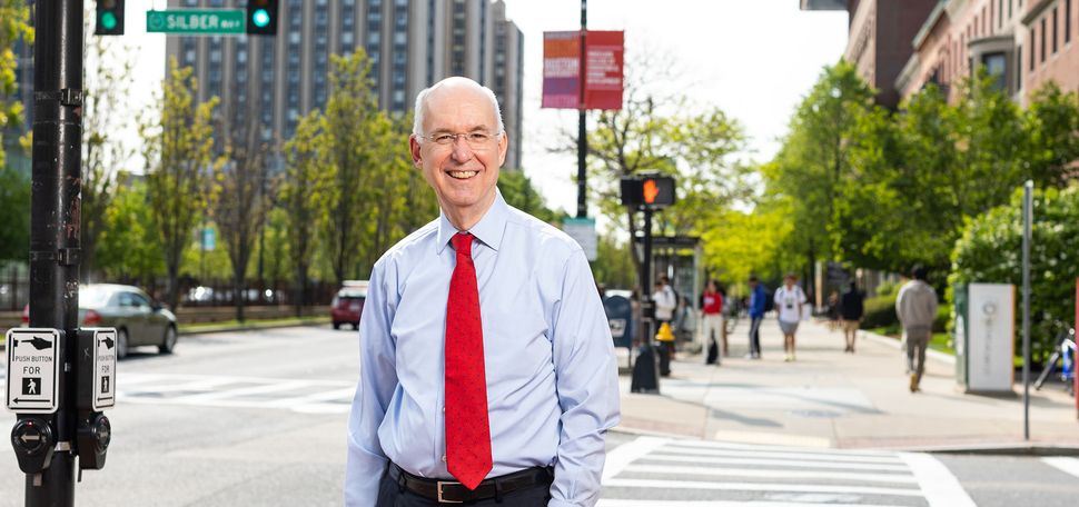 Photo: Kenneth Freeman, a white man wearing glasses, a light blue collared shirt, red tie, and black pants smiles and poses on a sidewalk on Commonwealth Avenue. Trees and various BU buildings can be seen in the blurry background on the sunny day.