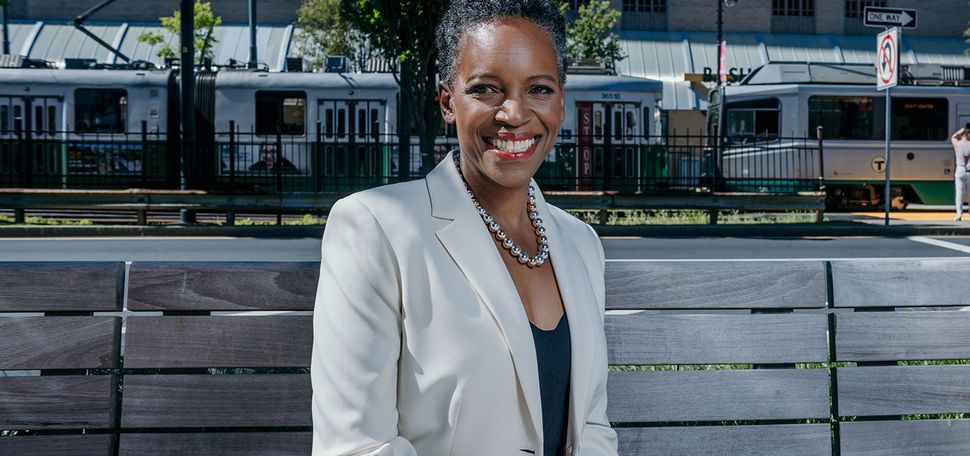 Photo: Boston University's new president Melissa Gilliam, a black woman with short dark hair in a formal outfit, sitting on a wooden bench and a smile in front of an MBTA train stop, in Boston on a sunny day.