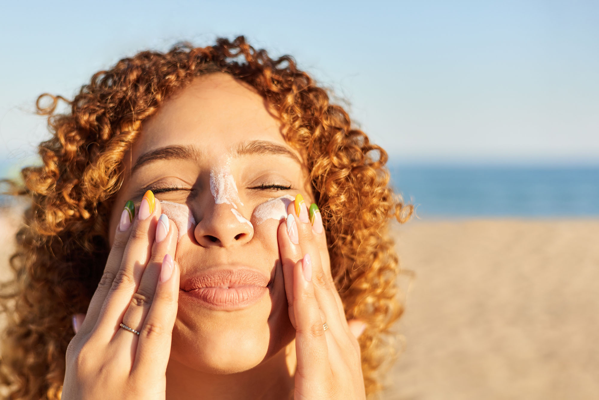 Photo: A picture of a smiling woman with curly hair applying sunscreen to her face on the beach