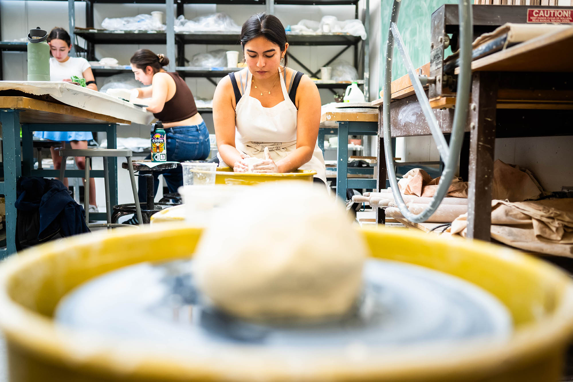 Photo: A young woman works at a potter's wheel as she preps to create a bowl. The mound of clay is in the foreground out of focus while she is in focus in the background.