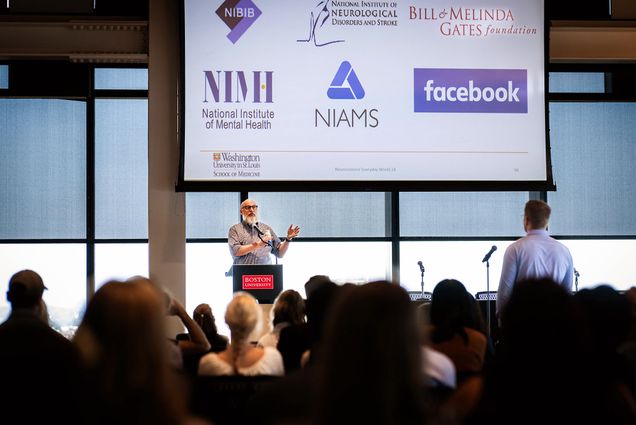 Photo: A man with a long white beard stands at a podium in front of a screen as he gives a speech to a small audience