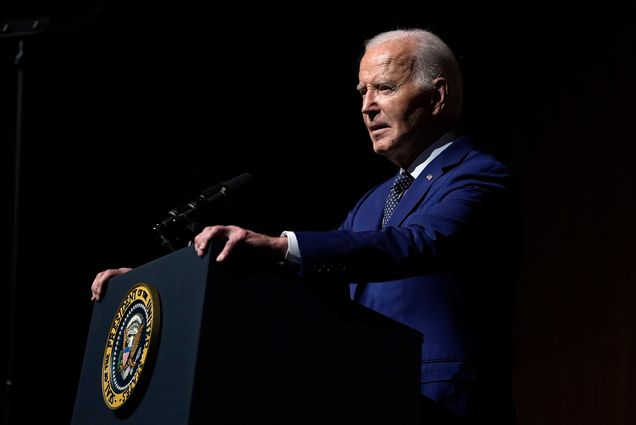 Photo: President Joe Biden speaks at an event. he stands at a podium as the right side of his face is illuminated.