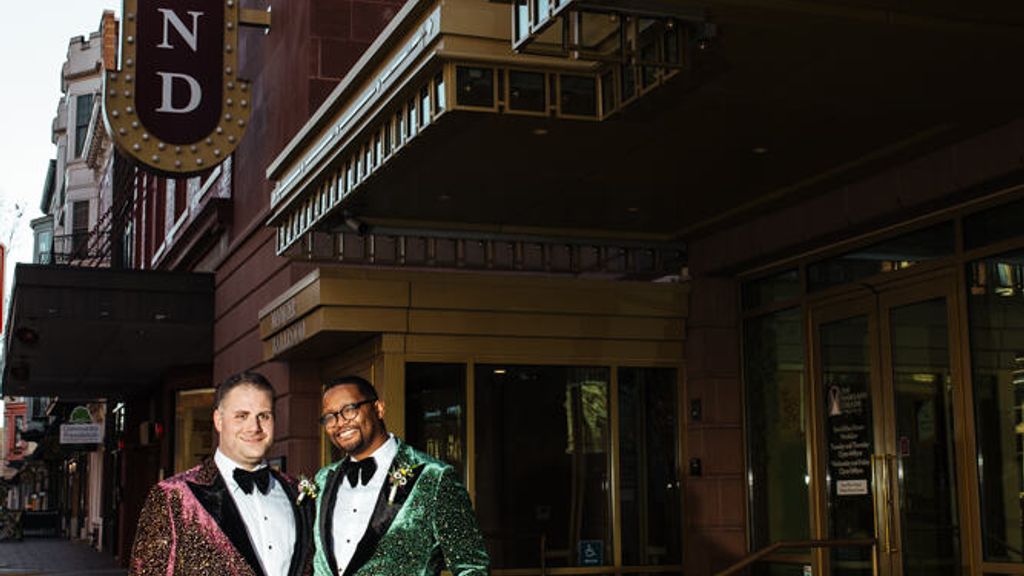 Photo: Two men in sparkly velvet suits embrace each other in front of a theatre