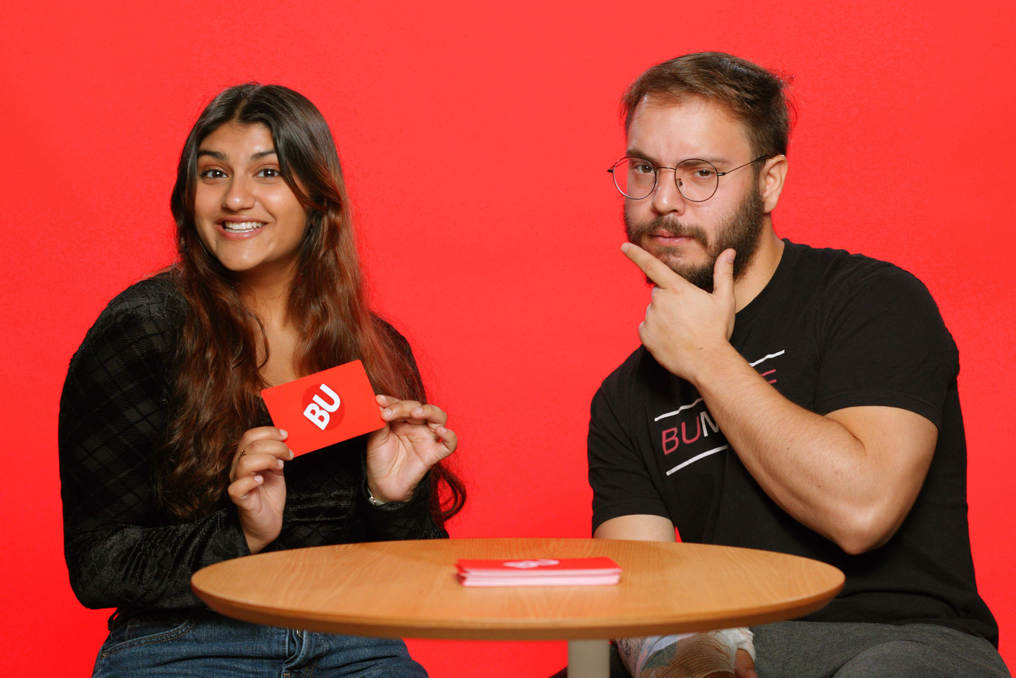 Photo: A picture of two students, a girl on the left and a boy on the right, posing in front of a red background. They are sitting at a table while the girl smiles and holds a red card that says "BU" and the boy places his pointer finger and thumb against his chin.