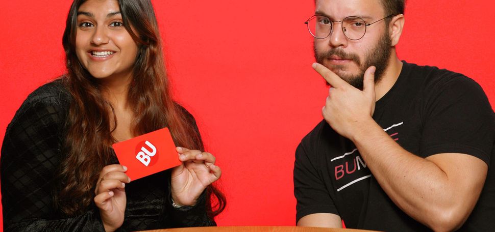 Photo: A picture of two students, a girl on the left and a boy on the right, posing in front of a red background. They are sitting at a table while the girl smiles and holds a red card that says "BU" and the boy places his pointer finger and thumb against his chin.