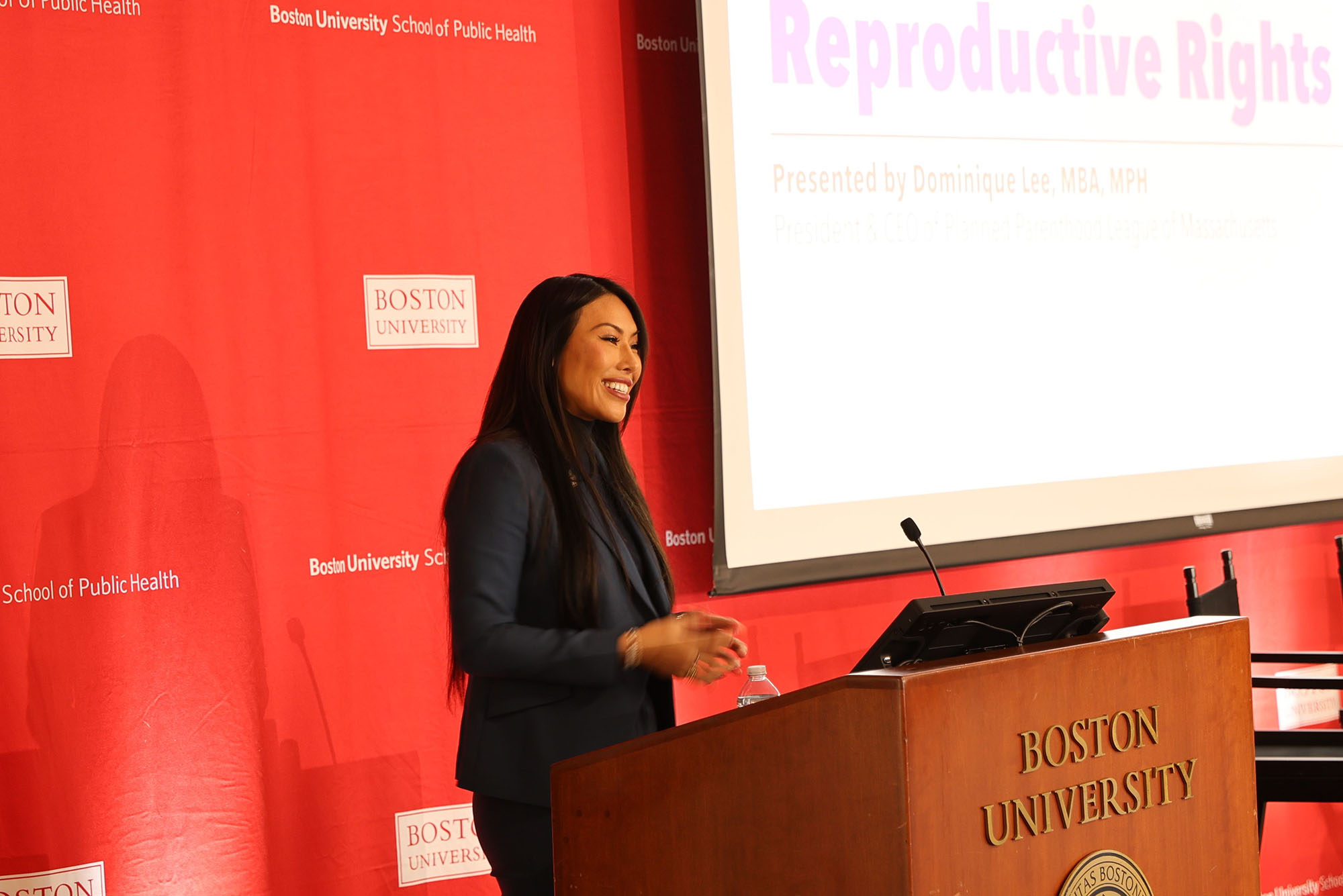Photo: A picture of a woman smiling and talking behind a podium. The wall behind her is red and has the "Boston University" logo many times