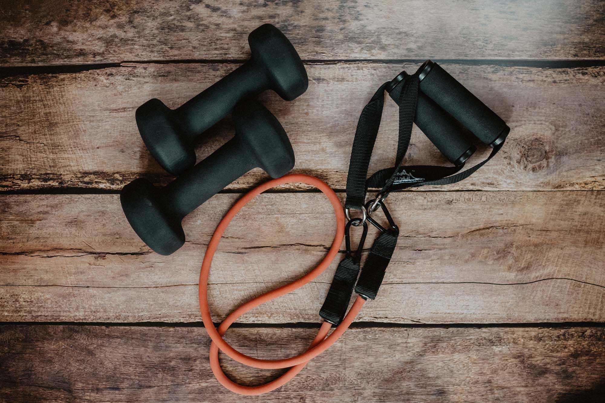 Photo: Overhead shot of 2 small black dumbbells and a black and red jump rope. Both are laid out on a wooden floor.