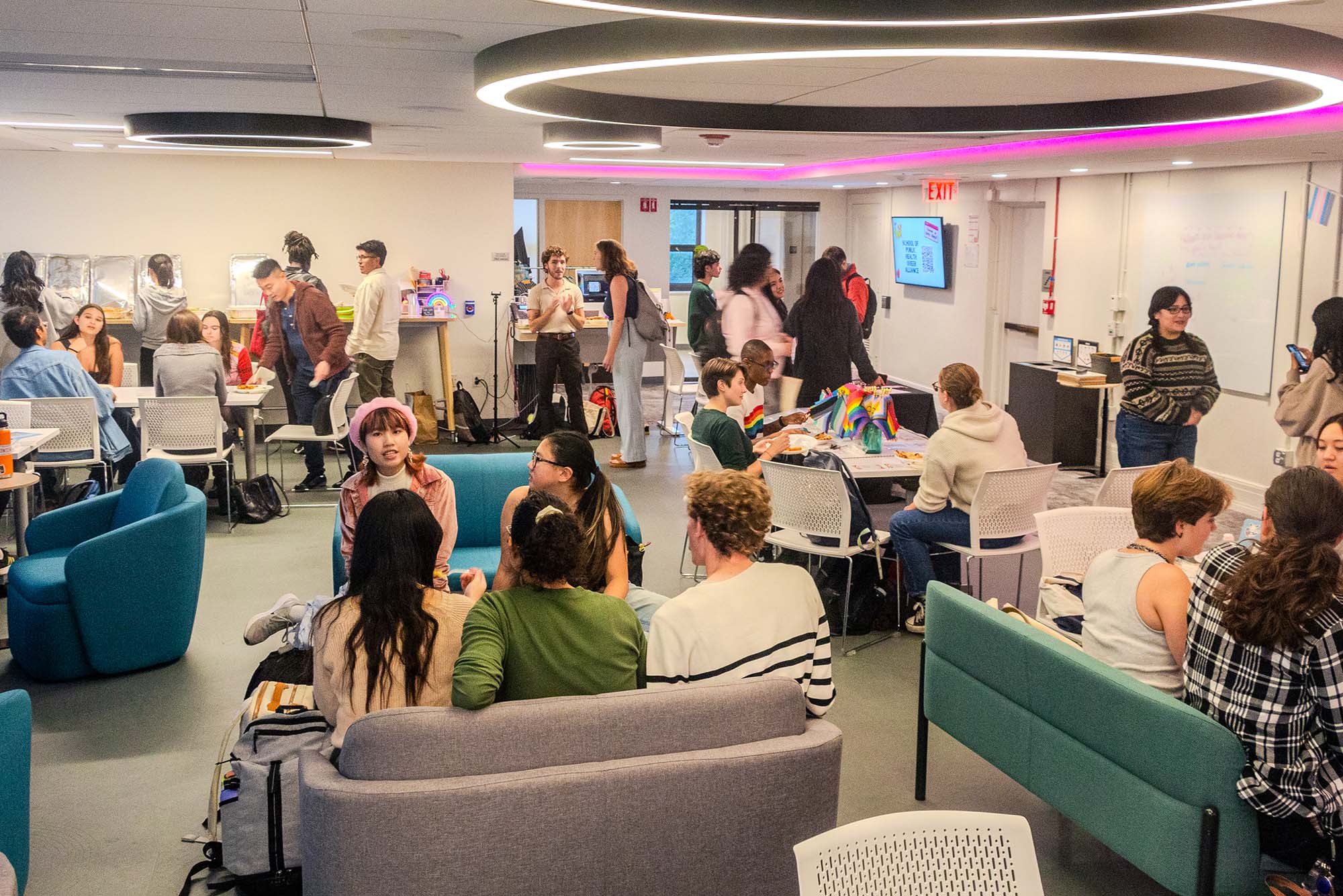 Photo: The LBGTQIA+ Student Resource Center with many people celerbating the kickoff of LGBTQIA+ History Month. There are several couches and arm chairs in the foreground that people are sitting at and chatting with one another, with others standing in the back by tables