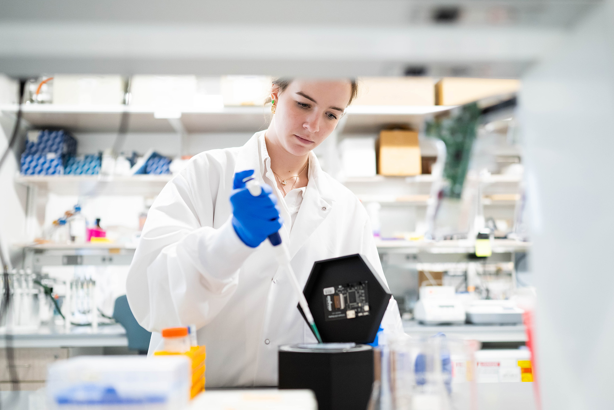 Photo: A picture of a female student in a lab coat and gloves demonstrating a soil monitoring device. The device is long with a pointed edge and is being placed into a black, hexagonal tube