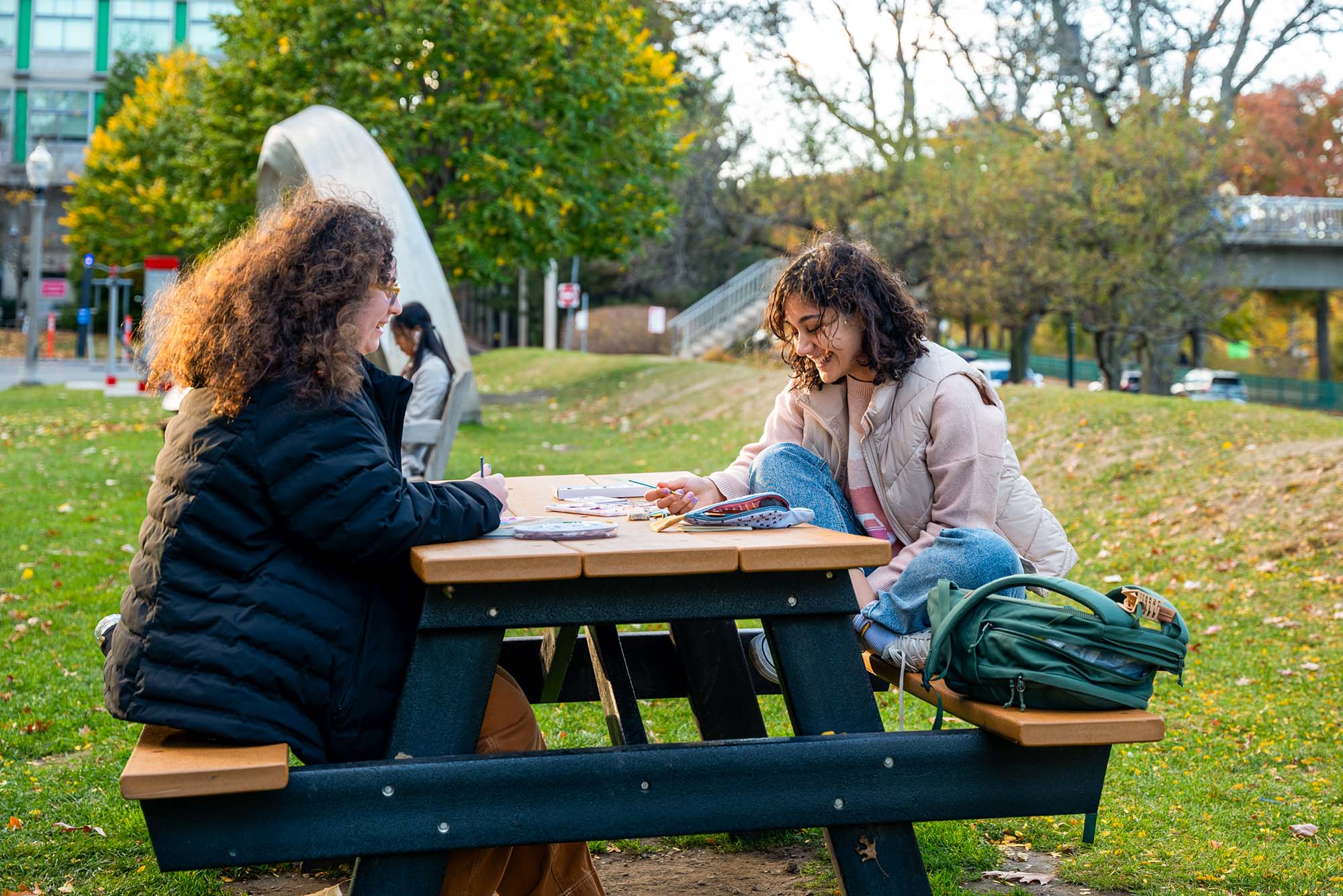 Photo: Two young women sit on a park bench, painting and enjoying the Autumn weather