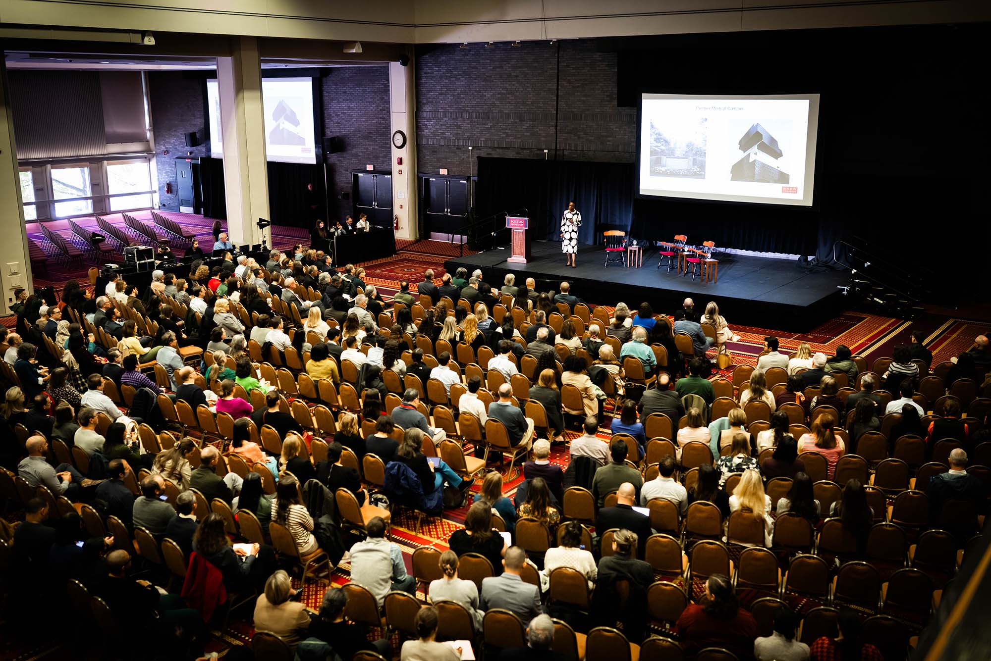 Photo: A wide shot of a town hall at Boston University with many people sitting in a crowd. BU President Dr. Melissa L. Gilliam is onstage in front of a projector screen