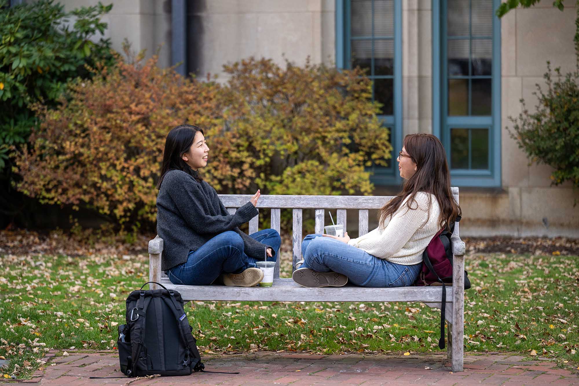 Photo: Two young women sitting cross legged on a park bench, chatting.