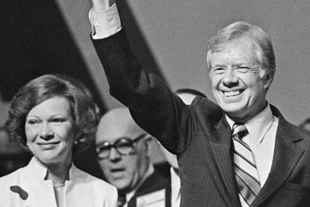 Photo: President Carter and Rosalynn Carter stand after President Carter gave a speech. He has his hand raised in a fist. They are smiling, him with his teeth and her with her mouth. It's a black and white photo.