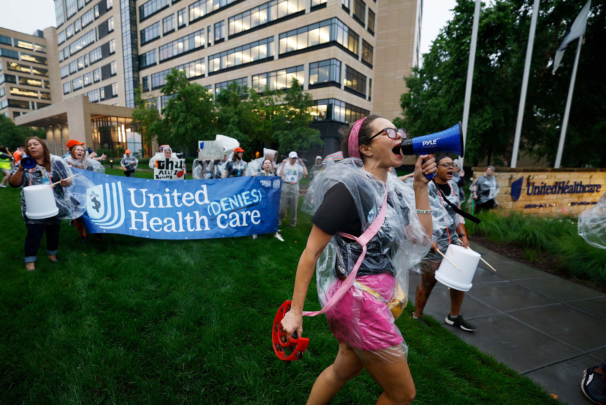 Photo: A protest against United Healthcare featuring a woman yelling into a megaphone and people holding a blue banner that says "United Denies Healthcare"