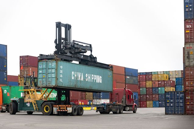 Photo: A cargo container labeled "China Shipping" being hoisted from a flatbad truck at a shipping port