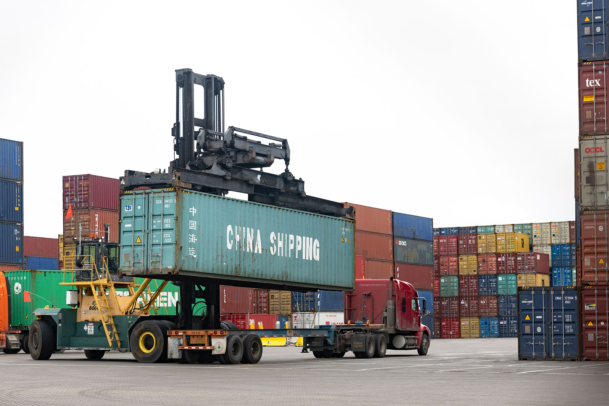 Photo: A cargo container labeled "China Shipping" being hoisted from a flatbad truck at a shipping port