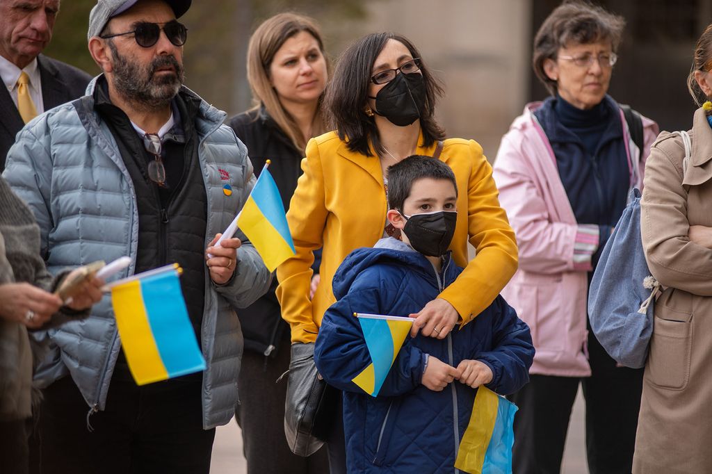 Assistant professor of physics and chemistry, Maria Kamenetska, stands in the center of the photo wearing a yellow puffer jacket holding her young son, Adam Liston, who is wearing a blue puffer coat. They are both holding a hand-held Ukrainian flag. The flag has a bold, light blue stripe on the top and and a bold, gold-yellow stripe on the bottom. On the left of the pair stands Kamenetska ’s father, Igor Tsinman. He is wearing a grey baseball cap that matches his grey puffer jacket which is overtop his black zip up. He also has sunglasses on and is carrying the same Ukrainian flag in his left hand. The group stands in front of two women--one in a black coat with light brown hair and the other in a light pink coat with darker and shorter brown hair. They also stand in front of a man in a black suit with a yellow tie, and just behind another woman in a camel coat with a jean draw-string bag. The people around the group are all holding the hand-held Ukrainian flag in their hands.