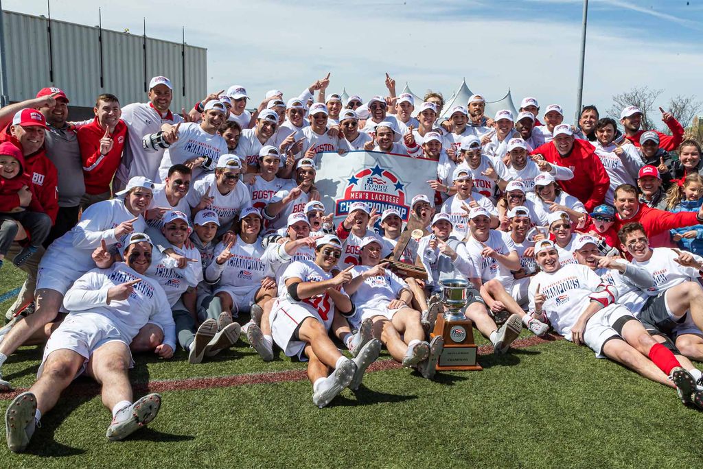 The entirety of the BU Lacrosse team, all of the men in their white uniforms, pose in front of and around a white banner titled "Men's Lacrosse Championship". Surrounding the team in various athleisure styles in the color of BU red are the coaches and supporters of the team. The entire group are seen with huge smiles and look very excited and happy.