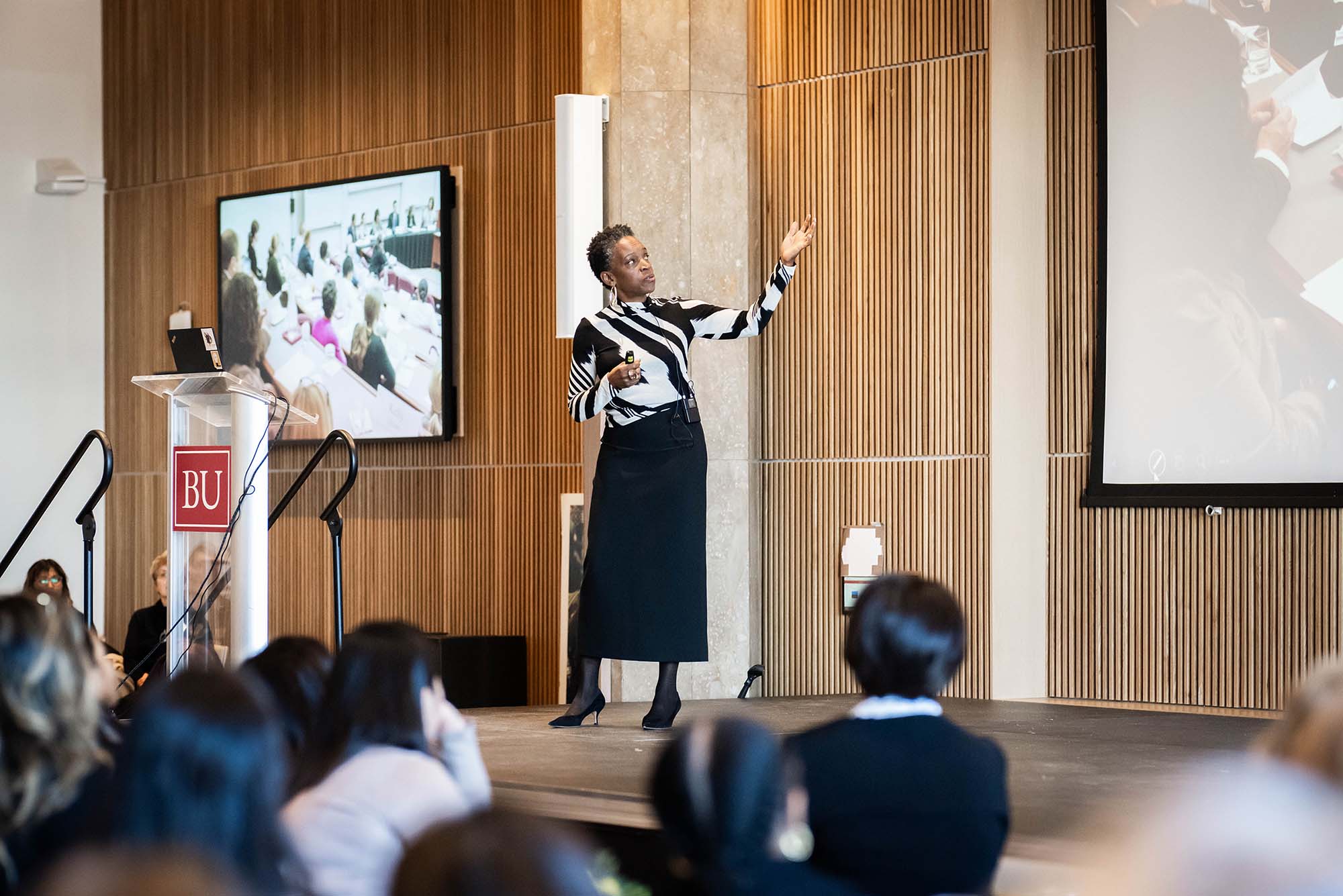 Photo: BU President Melissa Gilliam standing in front of a crowd and speaking. She is wearing a black and white striped shirt, long black skirt, tights, and black pointed-toe heels