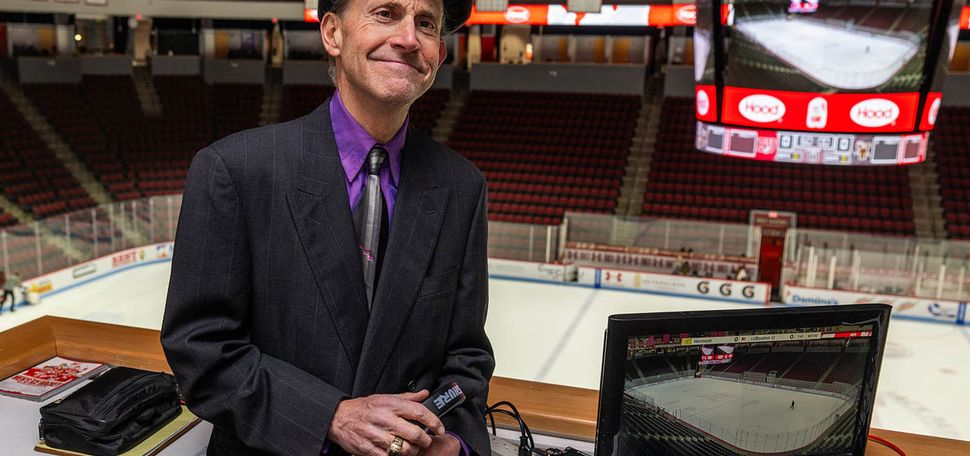 photo: A man wearing a striped black suit, purple shirt, black tie, and black bowler cap while leaning on an announcer's table at the BU hockey arena