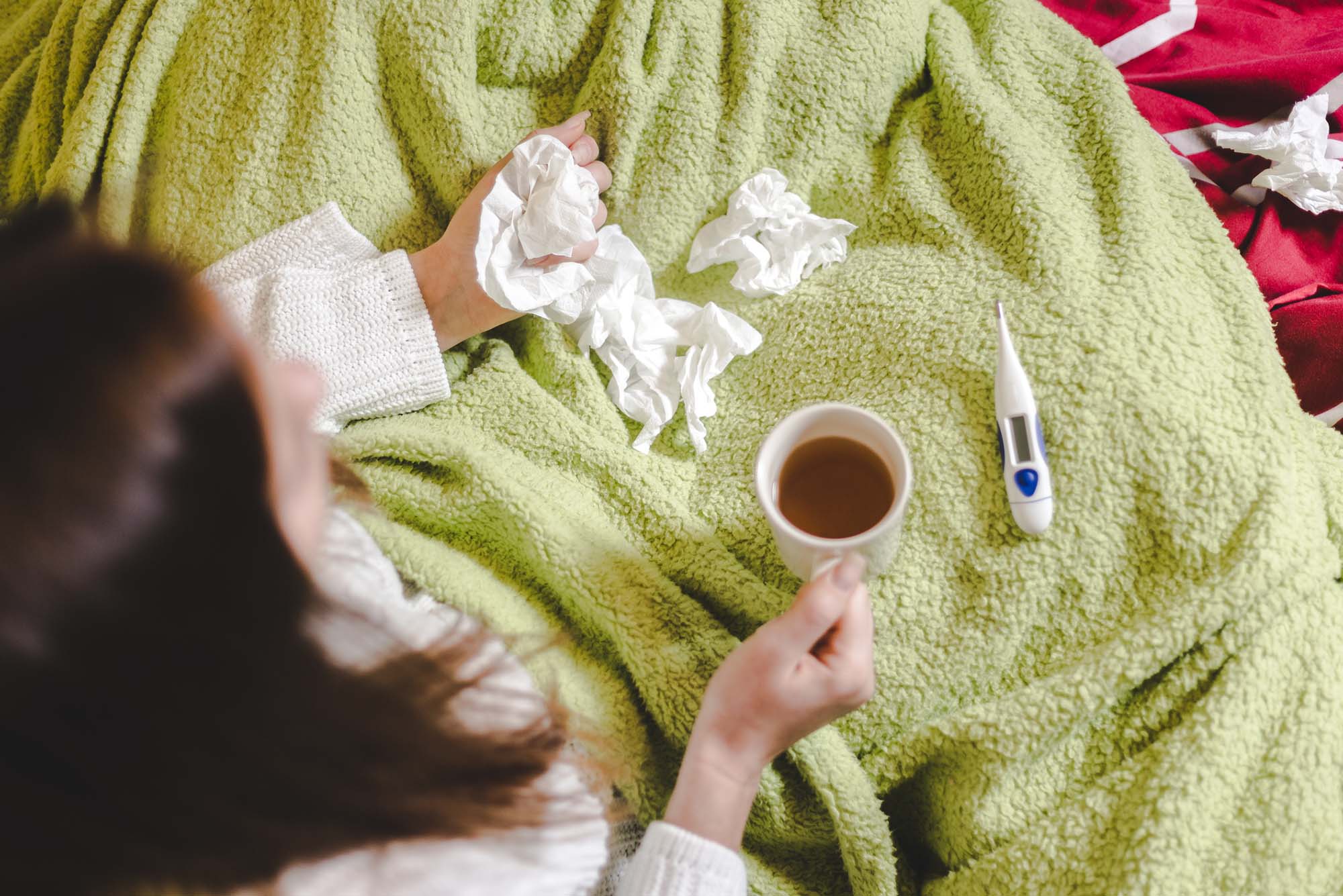 Photo: An above view of a woman sitting with a green blanket wrapped around her, tea in her hand, and crumpled up tissues strewn about the blanket