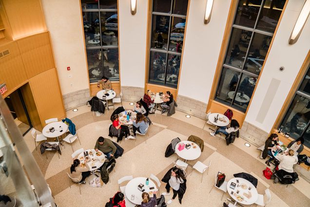 Photo: A birds-eye-view shot of the dining hall with ten round tables scattered about. All tables have some amount of various clothing and supplies on them. Some are void of people, the belongings there marking their space. Some tables host one, two, or three individuals eating, conversing, or preparing to leave the dining hall.