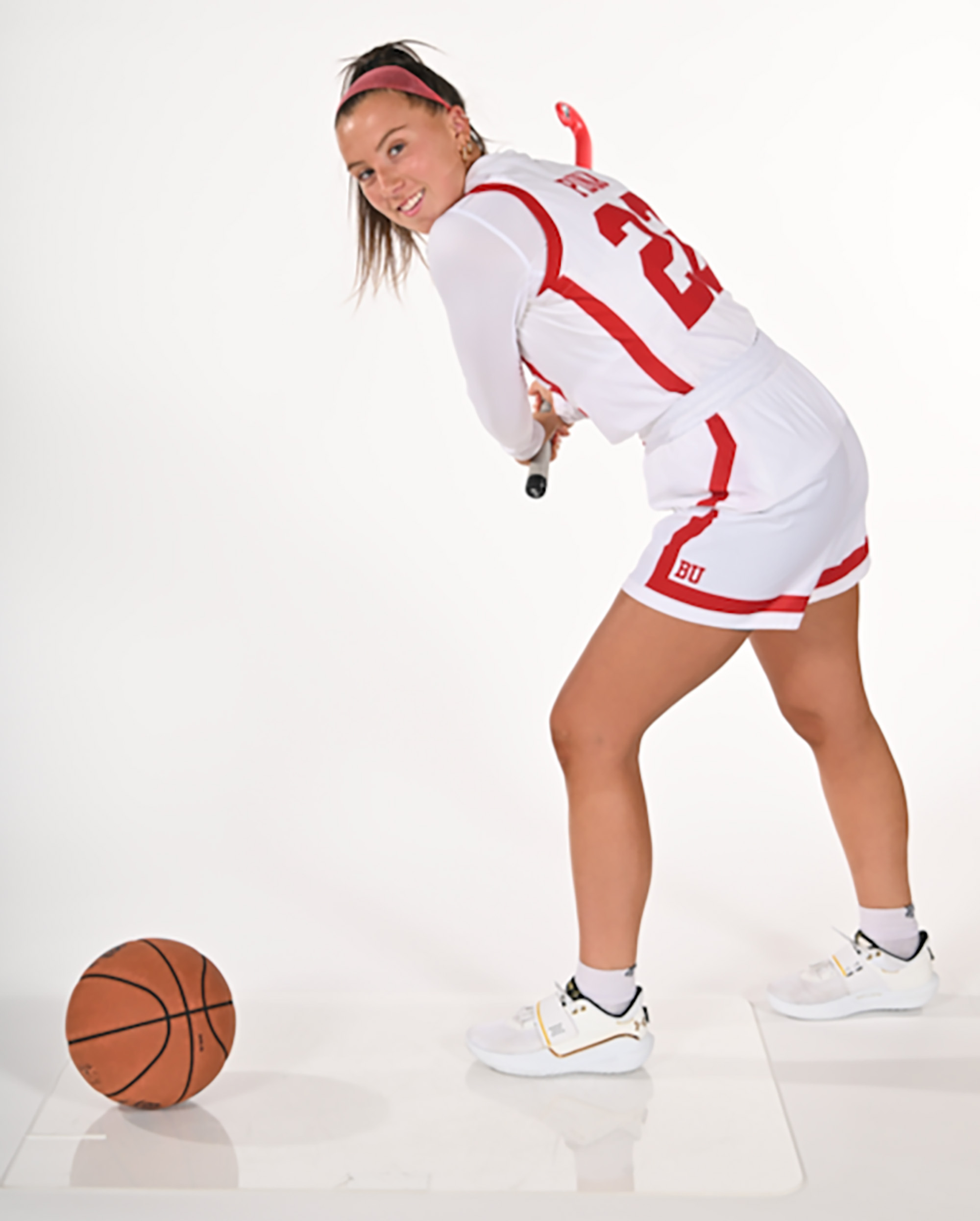 Maggie Pina (CAS’22, MET’24) tees up a basketball with her field hockey stick. She stands in the middle of a well-lit, all white background wearing a white uniform with red detailing. The basketball is to her left and she holds the field hockey stick in her right hand. She is also wearing flat, white sport shoes.