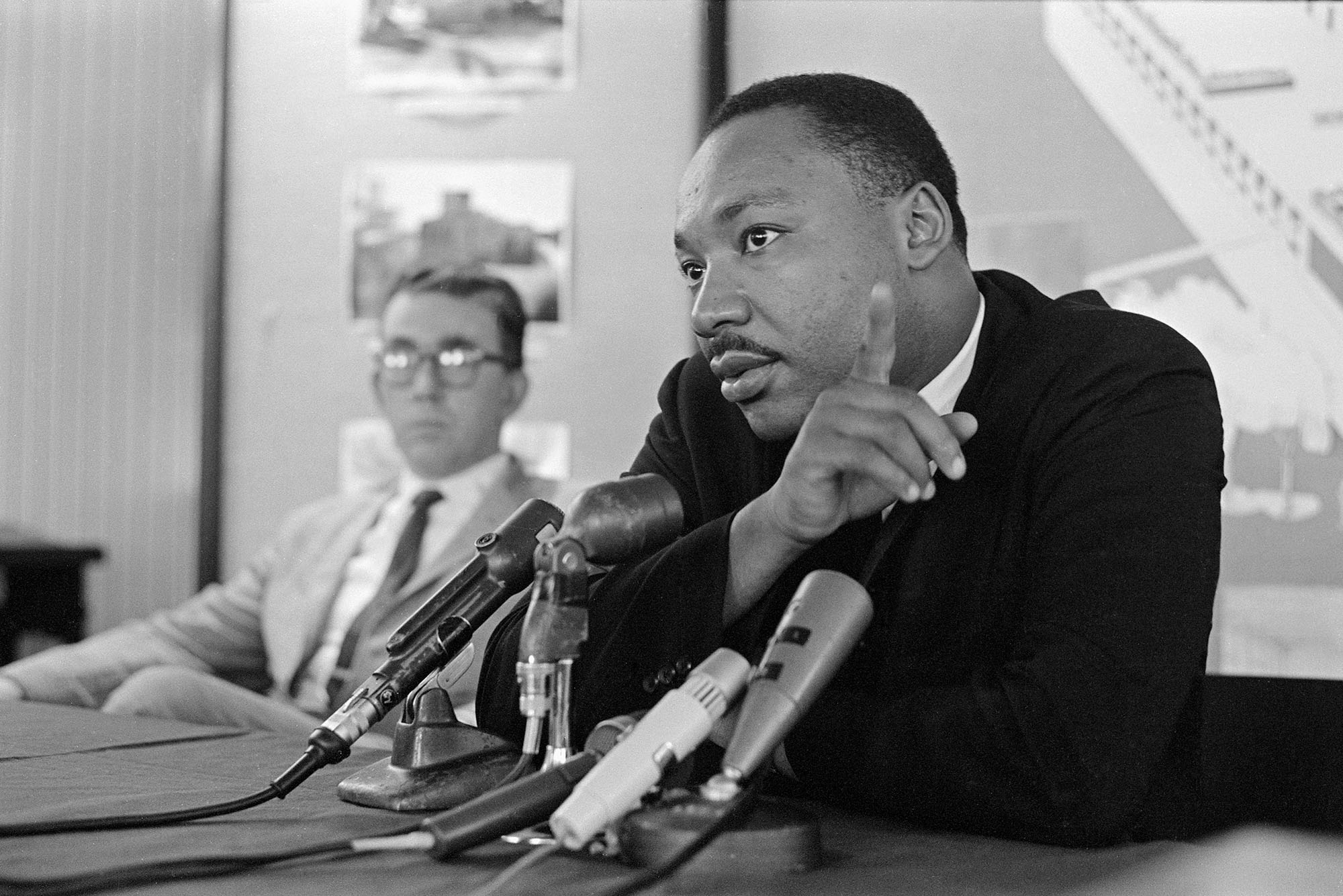 Photo: A black-and-white picture of Martin Luther King Jr. sitting down and speaking into a microphone with a man sitting to his left in the background