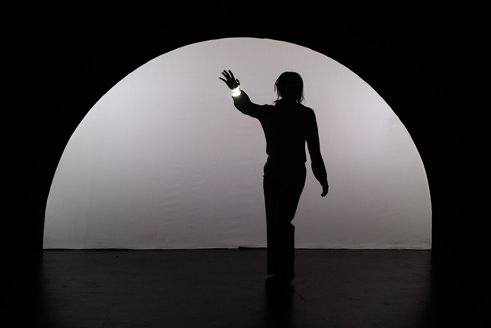 Photo: A person in shadow on a stage that has a white semi circle a backdrop and is otherwise dark. Their arm is extended towards the audience