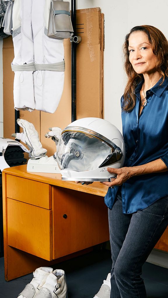 Photo: A woman leaning against a desk as she holds an astronaut helmet. The wall behind her has astronaut suits across it and a poster with an astronaut that says "Apply Now"
