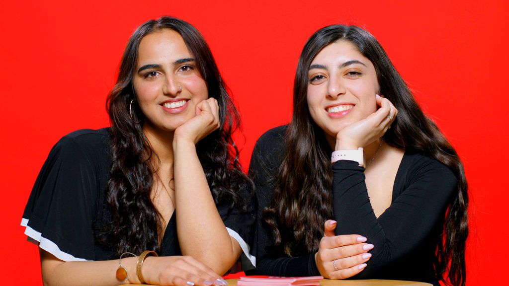Photo: Two girls smiling and posing with their head resting against their hand as they sit behind a table. There is a red background behind them