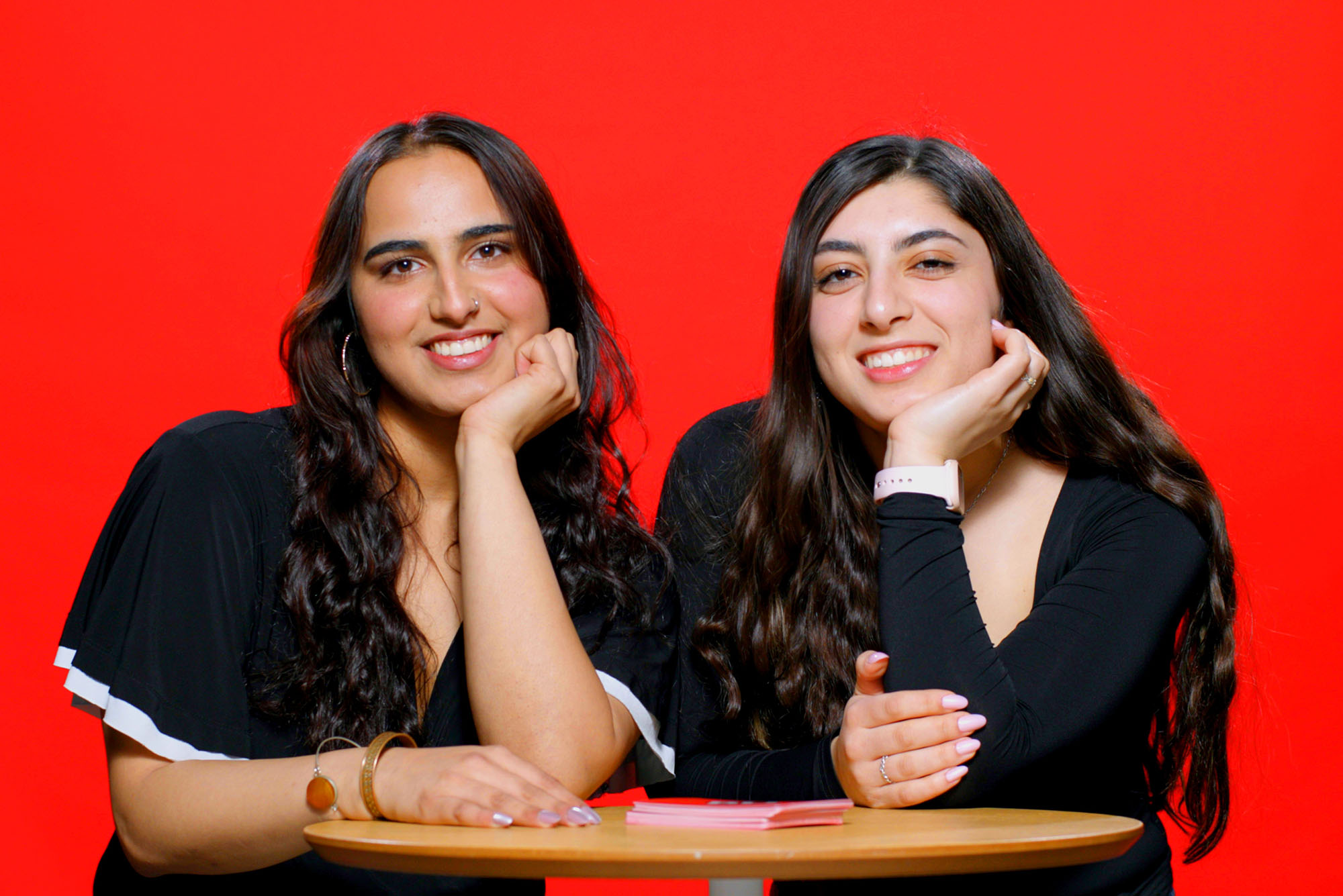 Photo: Two girls smiling and posing with their head resting against their hand as they sit behind a table. There is a red background behind them