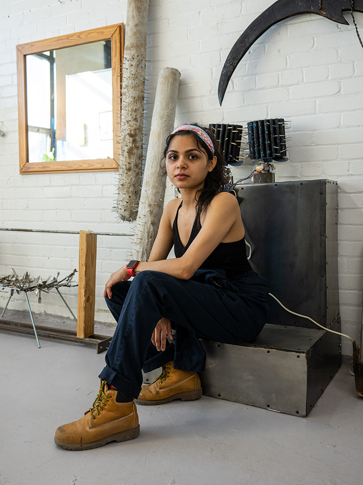 Photo: A woman with long dark hair wearing a bandana sits backwards on a chair inside of an art studio