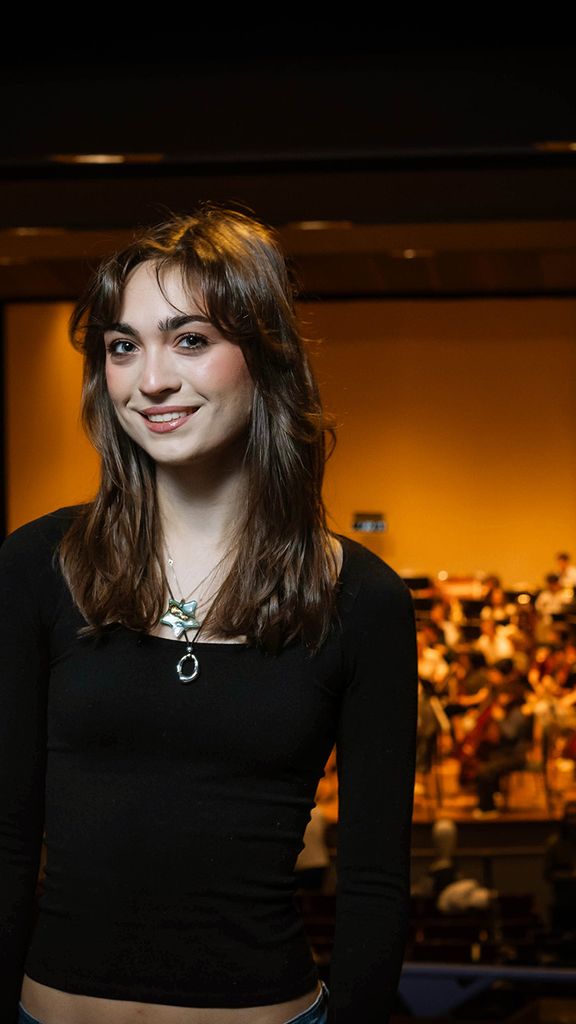 Photo: A girl with brown hair and bangs wearing a long sleeve black shirt and smiling in front of a stage